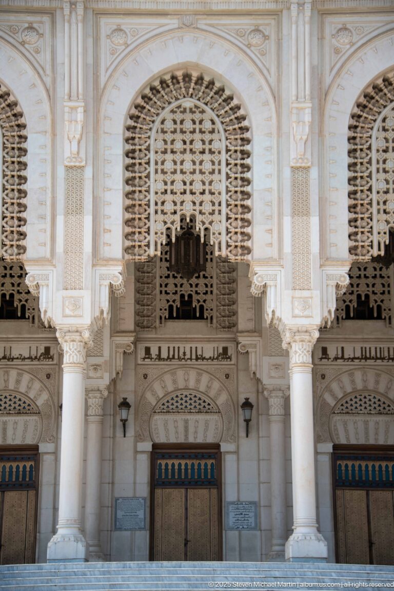 Close frontal view of ornate mosque entrance bays with white columns, pierced screens, carved arches, and dark doors.