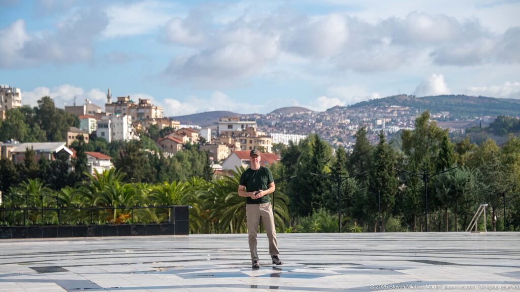 Person standing on the mosque’s broad patterned forecourt with trees, hills, and Constantine buildings in the distance.