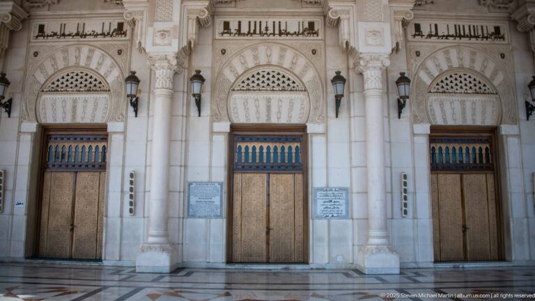 Three bronze-toned mosque doors beneath carved arches, Arabic calligraphy bands, and dark wall lanterns.