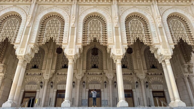 Low-angle view through a tall carved entrance arcade with columns, pierced screens, doors, and a person for scale.