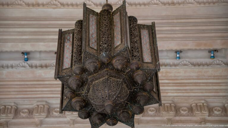 Large faceted hanging lantern of pierced dark metal suspended beneath a carved pale stone ceiling.