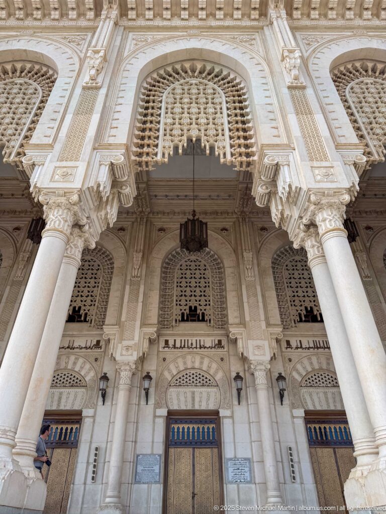 Upward view of a mosque entrance bay with large columns, carved arch, pierced screen, lamps, and metal door.
