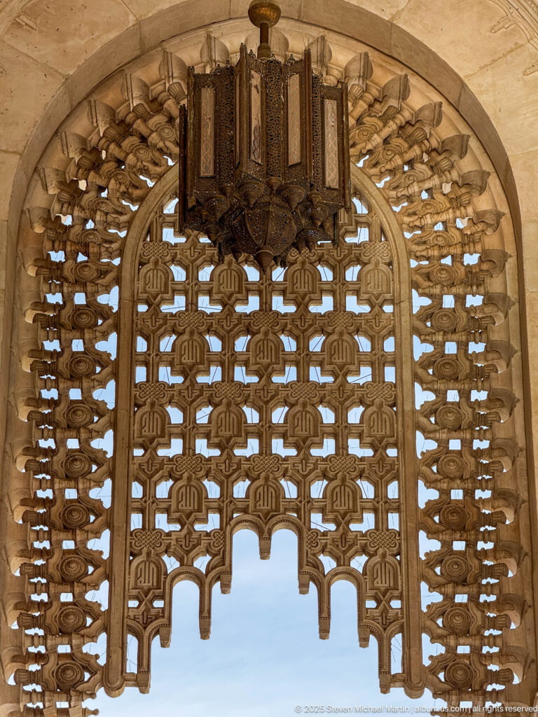 Arched pierced screen with repeating calligraphic geometric motifs and a dark hanging lantern above blue sky.