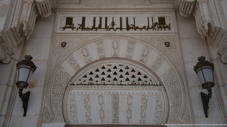 Carved mosque entrance arch with Arabic inscription above, triangular perforations, and wall lanterns on both sides.