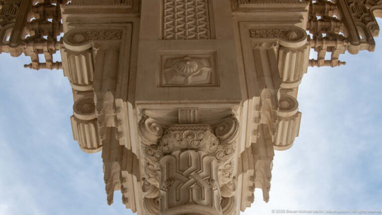 Close upward view of an elaborately carved pale stone capital and projecting bracket against blue sky.