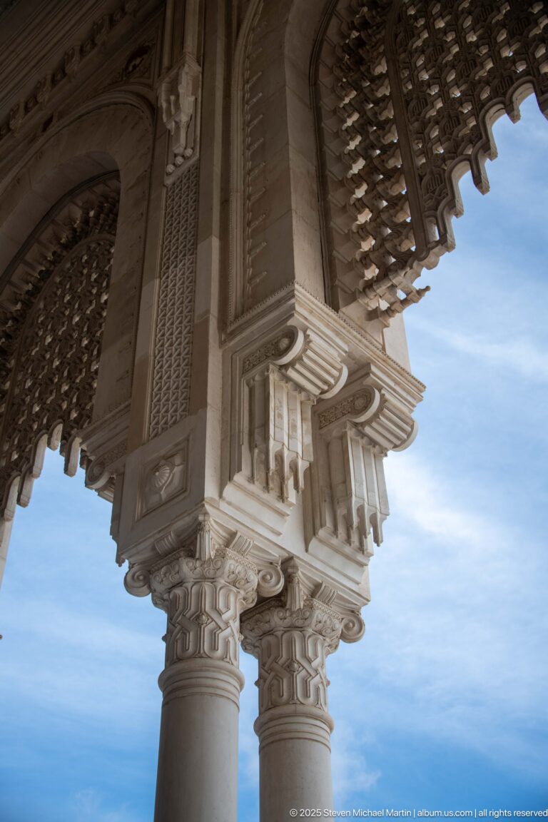 Low-angle detail of a mosque column meeting an ornate carved bracket and beam beneath an openwork canopy.