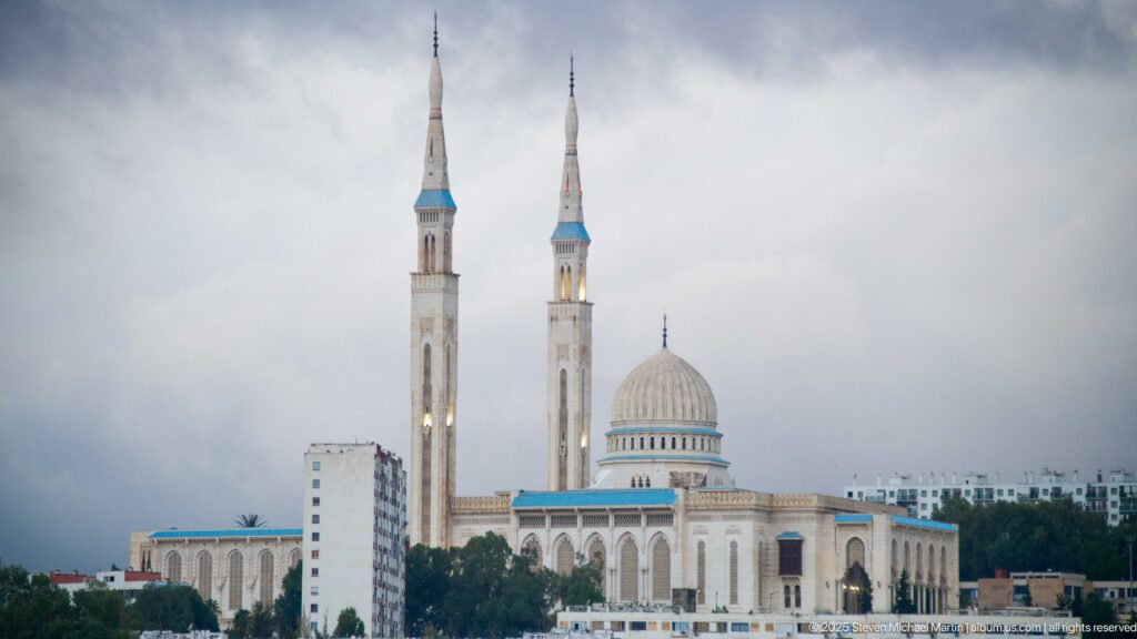 Distant daytime view of Amir Abdel Kader Mosque with twin minarets, ribbed dome, trees, and nearby buildings.