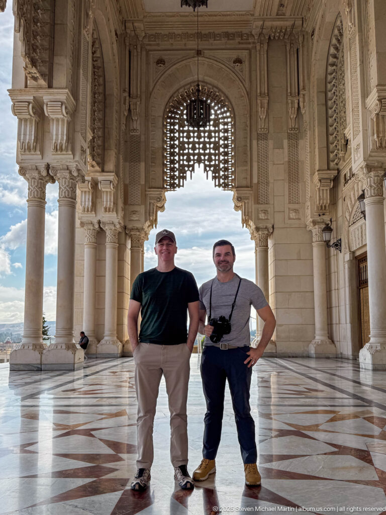 Two men standing beneath a monumental mosque portico with columns, carved arches, hanging lamps, and patterned floor.