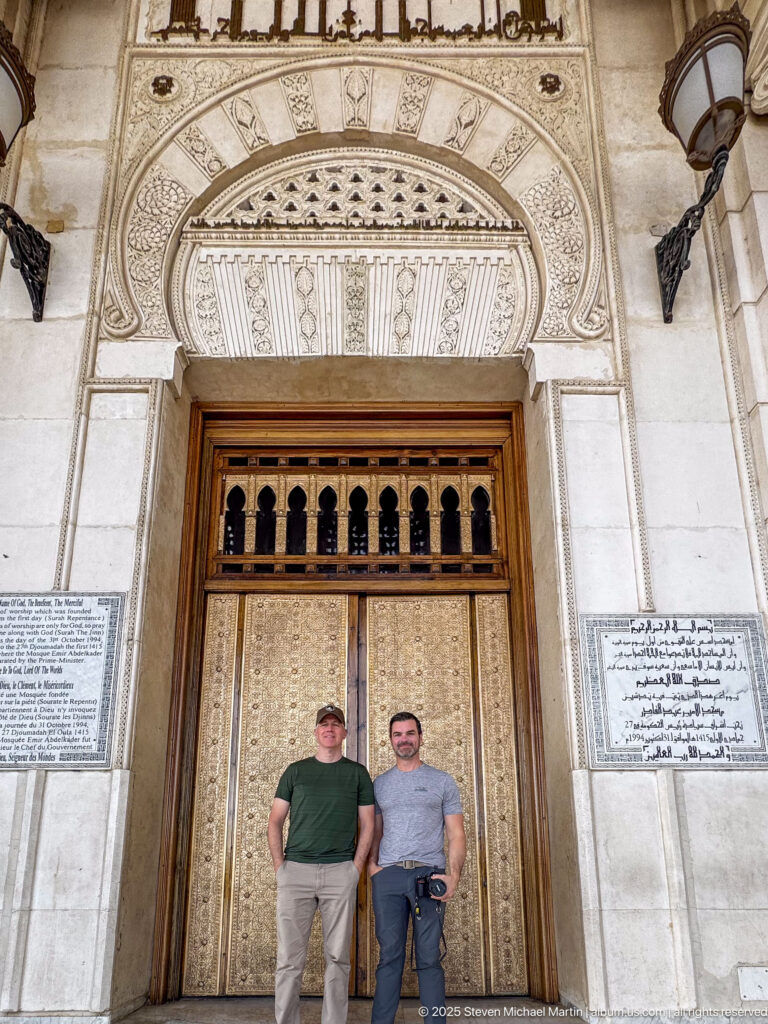 Two men standing before a tall patterned metal mosque door framed by carved stone and inscription plaques.