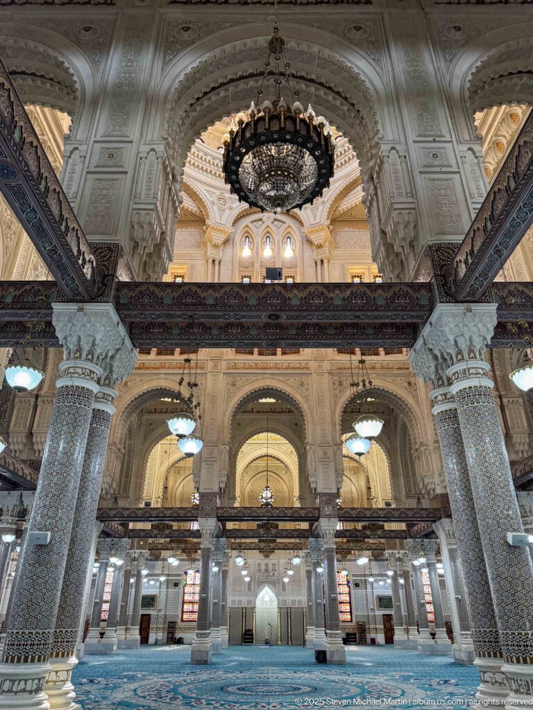 Grand mosque prayer hall with blue carpet, mosaic-covered columns, upper gallery, carved arches, and large chandelier.