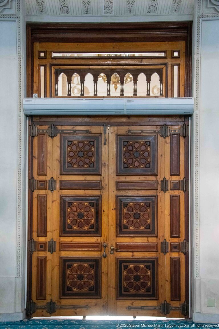 Large wooden double door with geometric inset panels and a glazed transom of small rounded arches.
