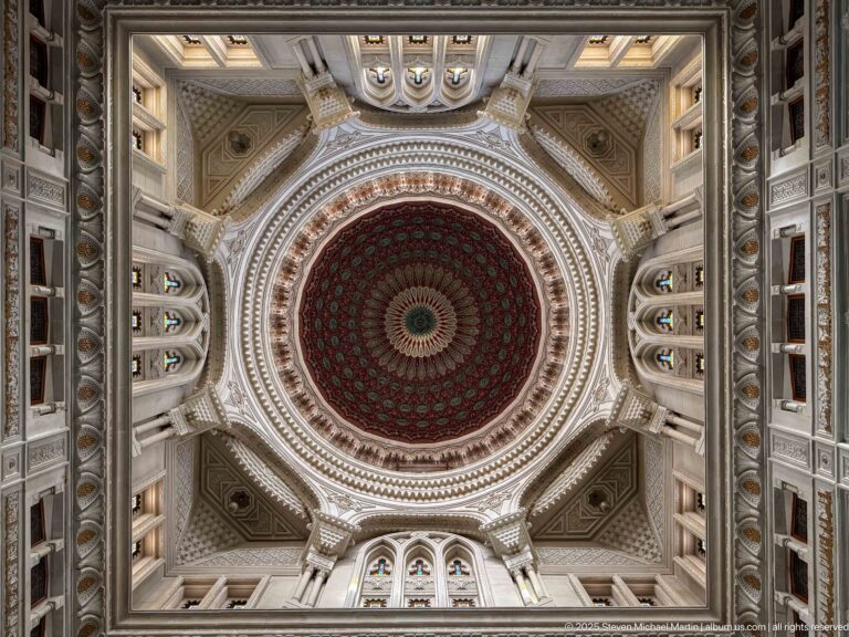 Upward view of an ornate circular mosque dome with concentric red and green patterning inside a carved square surround.