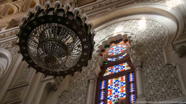 Dark circular chandelier hanging beside a brightly colored stained-glass window in an ornate carved mosque interior.