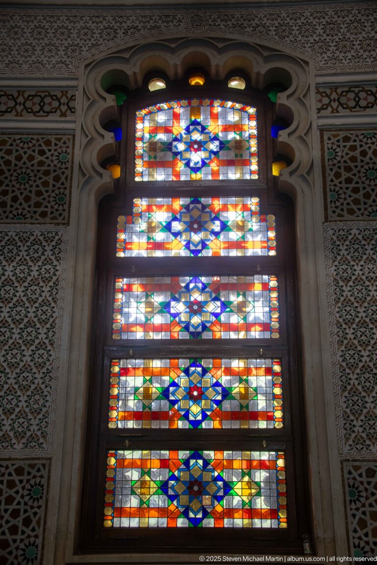 Tall multicolored stained-glass window with stacked geometric patterns framed by intricately decorated wall panels.