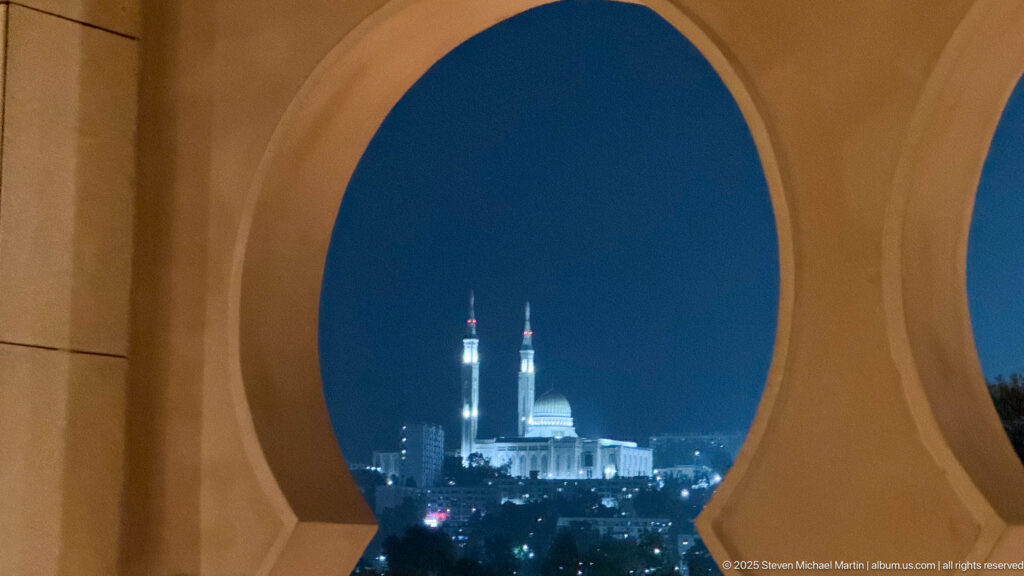 Illuminated Amir Abdel Kader Mosque seen at night through a large ochre architectural arch.