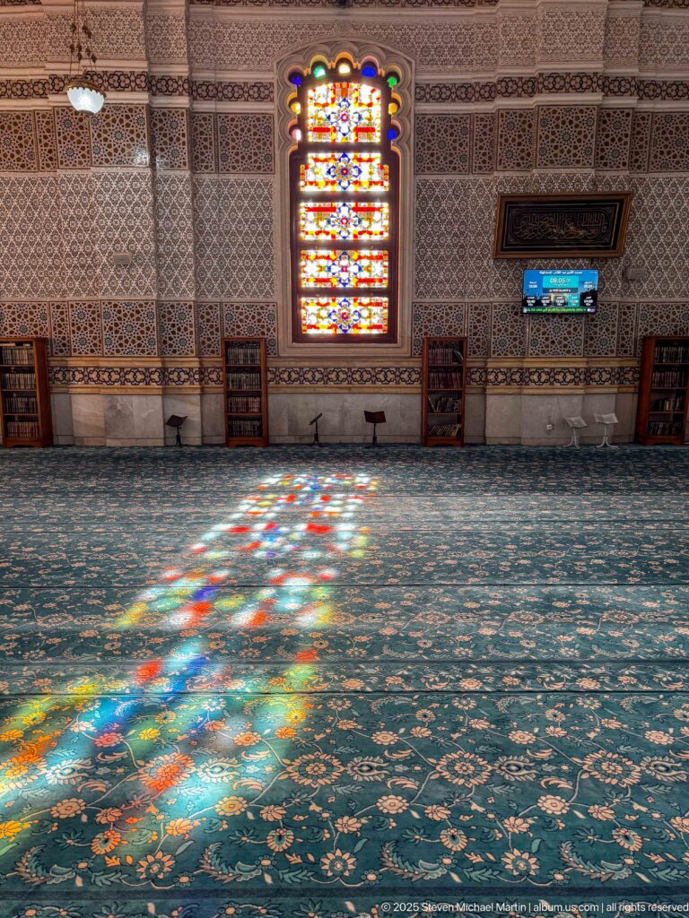 Multicolored sunlight from a stained-glass window spreading across blue mosque carpet beneath a patterned interior wall.