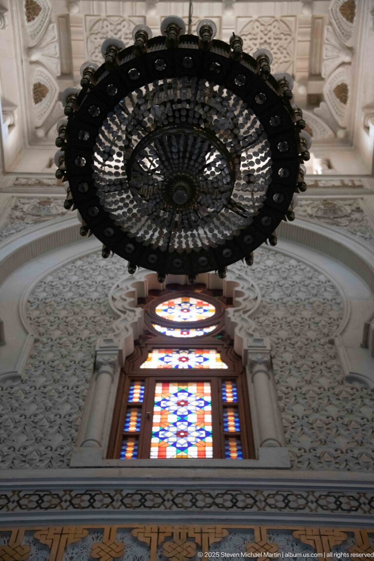 Large circular chandelier above a colorful stained-glass window and carved pale wall decoration.
