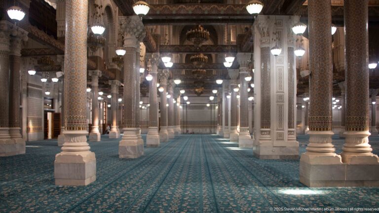 Long central aisle through the mosque prayer hall with blue carpet, decorated columns, hanging lights, and chandeliers.