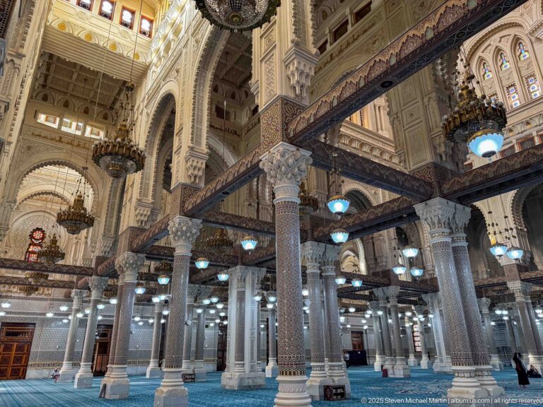 Oblique view across a blue-carpeted mosque hall with mosaic columns, decorated beams, hanging lamps, galleries, and high arches.