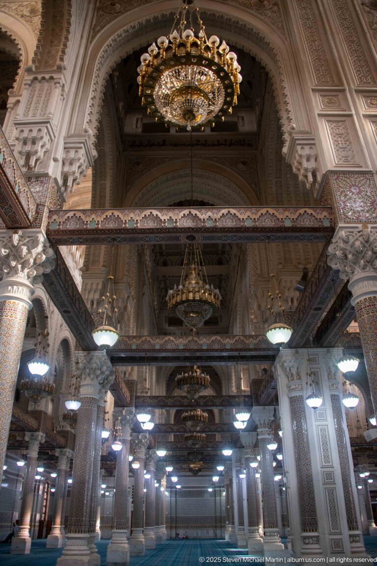 Rows of chandeliers and decorated beams beneath the mosque’s high vaulted interior, with patterned columns below.