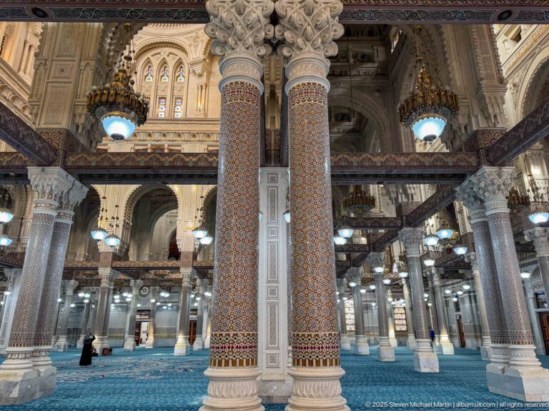 Pair of tall mosaic-covered columns with ornate carved capitals inside the blue-carpeted mosque prayer hall.