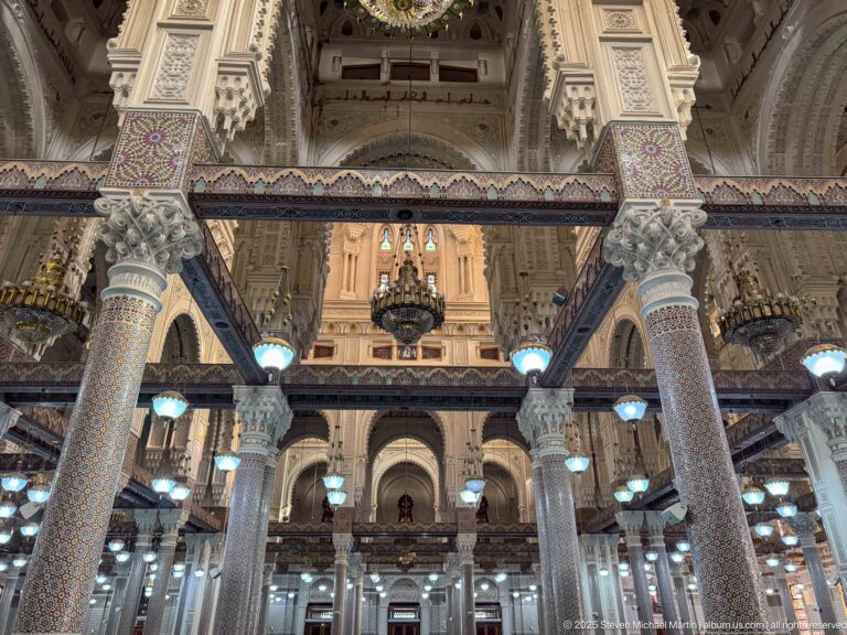Interior view framed by mosaic columns, with decorated beams, chandeliers, high arches, galleries, and blue carpet beyond.