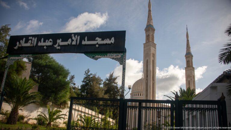 Arabic entrance sign and gate in the foreground with the mosque’s dome and twin minarets beyond.