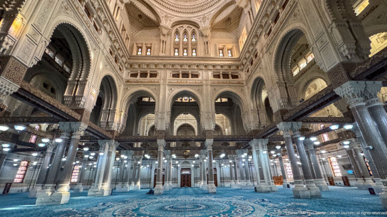 Symmetrical mosque interior with blue carpet, rows of columns, decorated beams, high arches, upper windows, and central carpet medallion.