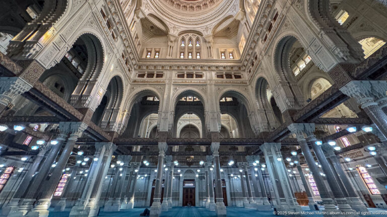 Symmetrical mosque prayer hall with blue carpet, rows of decorated columns, dark beams, high arches, galleries, and upper windows.