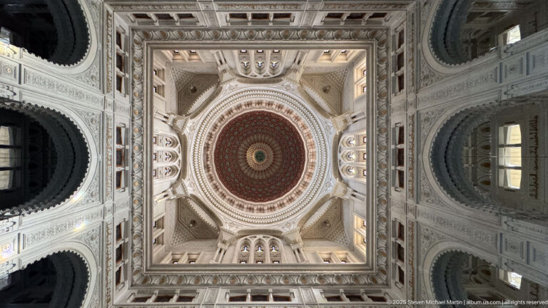 Upward view of a richly patterned circular mosque dome centered within carved square framing, arches, and corner vaults.