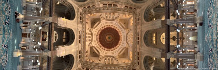 Rotated axial view through the mosque prayer hall toward a large patterned dome, flanked by columns, galleries, arches, and lights.