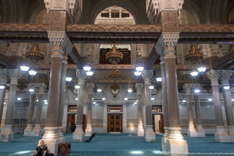 Blue-carpeted mosque hall with patterned columns, decorated beams, chandeliers, wooden doors, and a few seated people.