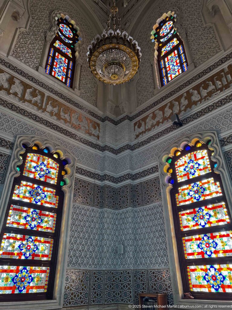 Two brightly colored stained-glass windows meeting at a decorated mosque corner beneath a hanging chandelier and smaller upper windows.