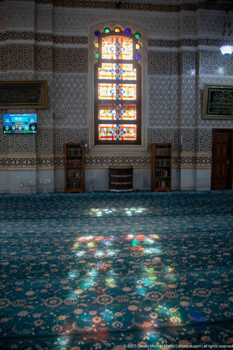 Stained-glass window above patterned mosque carpet, with colored reflections, shelving, framed panels, and low wooden furnishings.