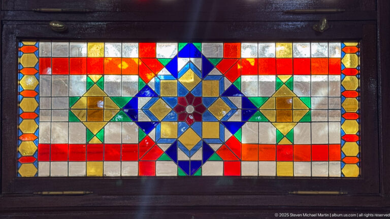 Geometric stained-glass panel in a dark wooden frame, composed of red, blue, green, amber, and clear polygonal panes.