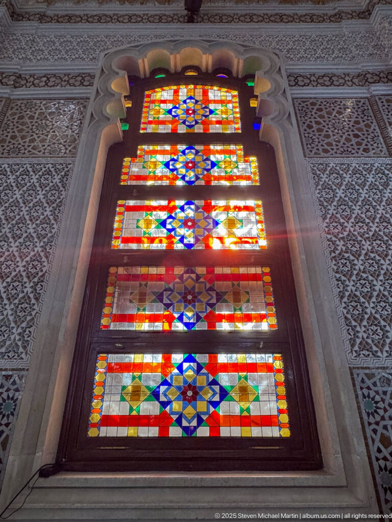 Tall arched stained-glass window with stacked multicolored geometric panels surrounded by pale carved and patterned wall decoration.