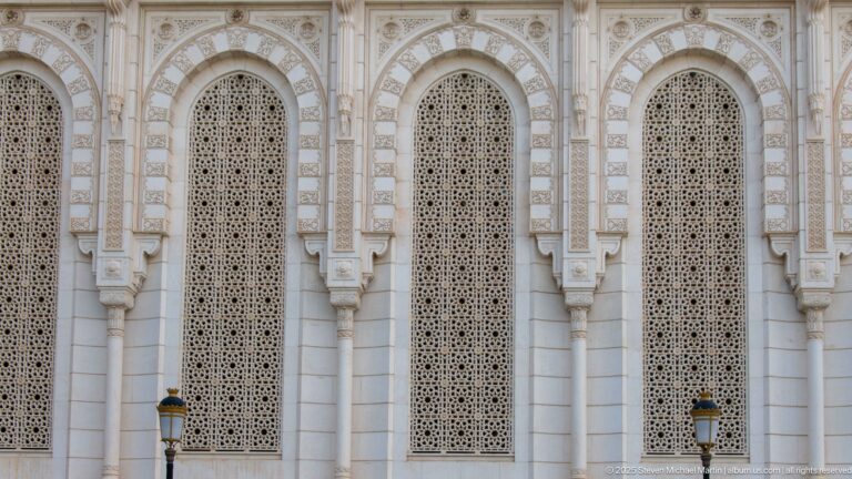 Three tall arched windows with intricate geometric screens and carved white surrounds on the mosque façade.