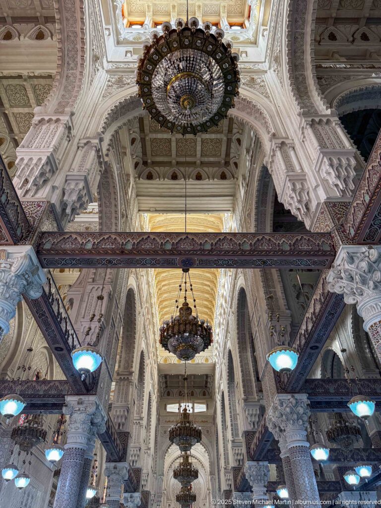 Axial interior view beneath large chandeliers, with carved arches, decorated columns, and repeating bays extending through the prayer hall.