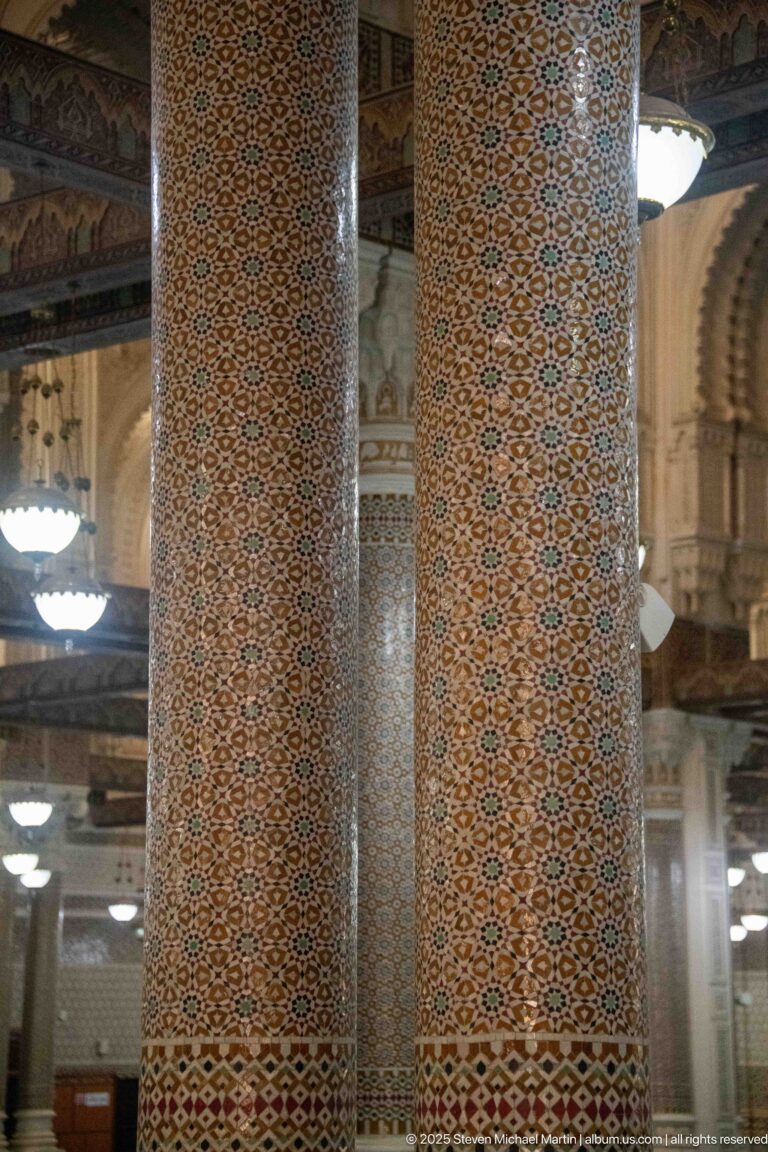 Two large interior columns covered in dense repeating geometric patterns, with arches and lights of the prayer hall visible behind them.