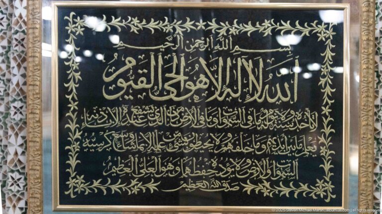 Framed black panel with large gold Arabic calligraphy, smaller lines of script, and a gold leafy border.