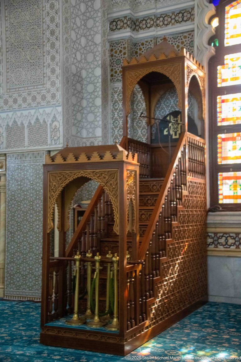 Carved wooden minbar with stairs, balustrade, and canopied platform beside a brightly colored stained-glass window.