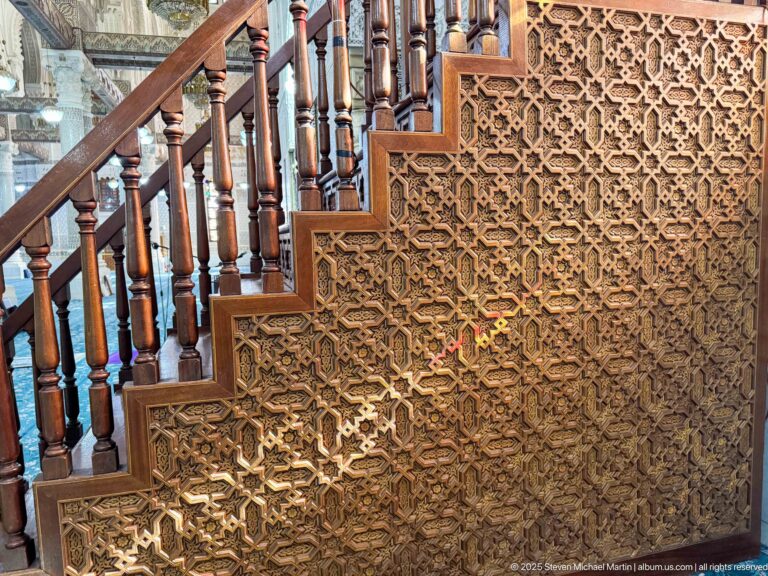 Close view of a minbar staircase with turned wooden balusters and an intricately carved panel of interlocking star patterns.