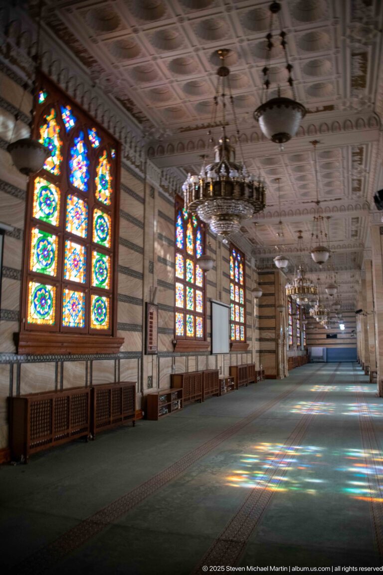 Long mosque aisle with stained-glass windows, hanging chandeliers, striped walls, and colored light falling across green carpet.