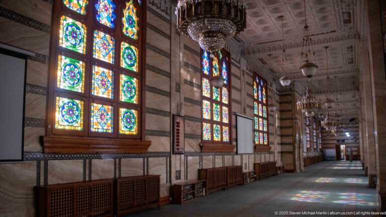 Stained-glass windows and chandeliers repeat along a striped interior wall beside the mosque’s carpeted prayer space.