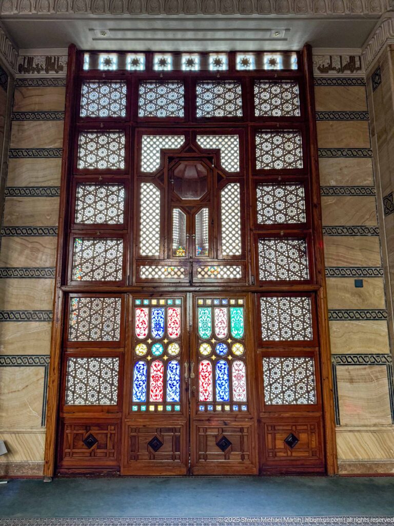 Large symmetrical wooden doorway with lattice panels, geometric carving, a projecting central feature, and small colored-glass panes.