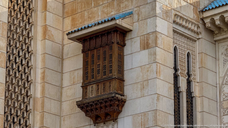 Projecting carved wooden window with lattice panels and brackets set into the mosque’s pale stone wall.