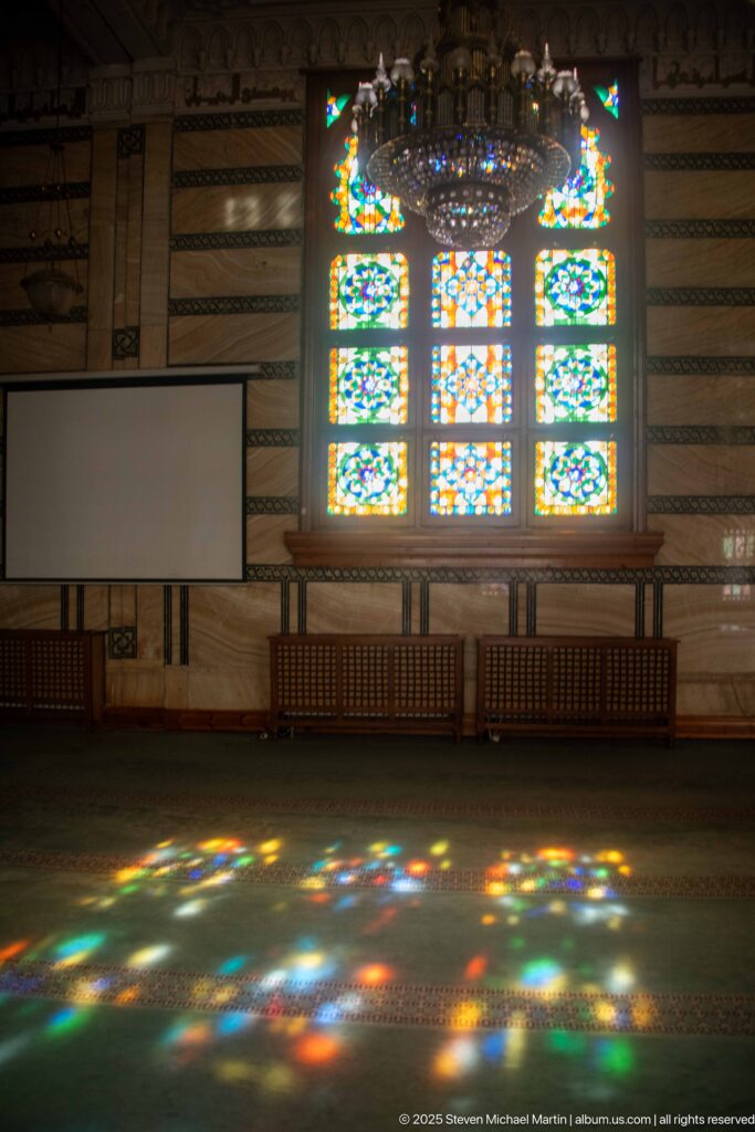 Multicolored stained-glass reflections spread across green mosque carpet beneath a chandelier and bright geometric window.