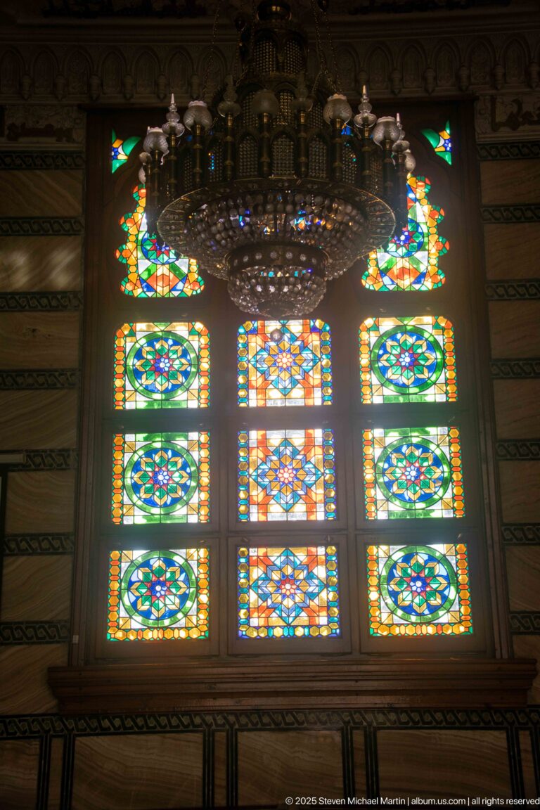 Large chandelier hanging before a nine-panel stained-glass window with vivid green, blue, yellow, and red geometric designs.