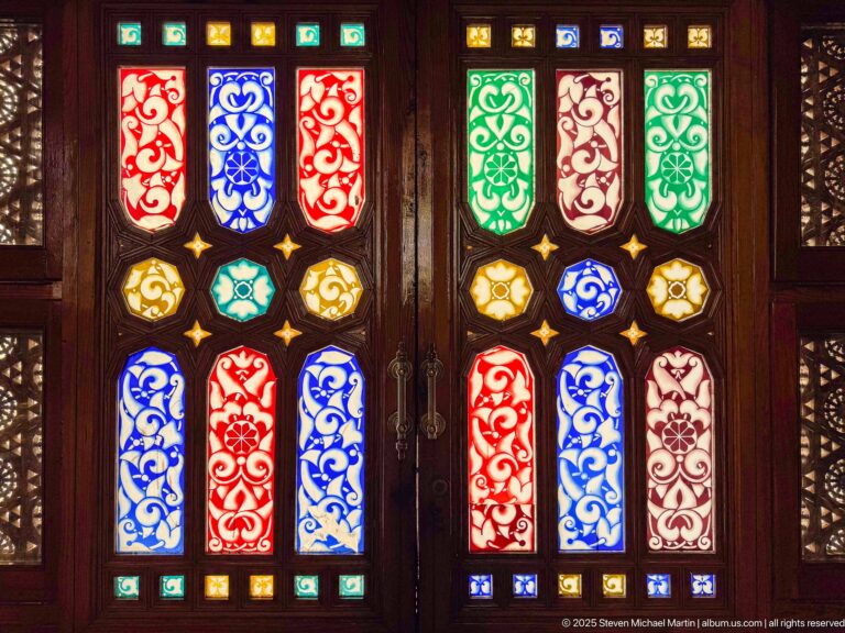 Double wooden doors filled with red, blue, green, amber, and white stained-glass panels arranged in symmetrical ornamental patterns.