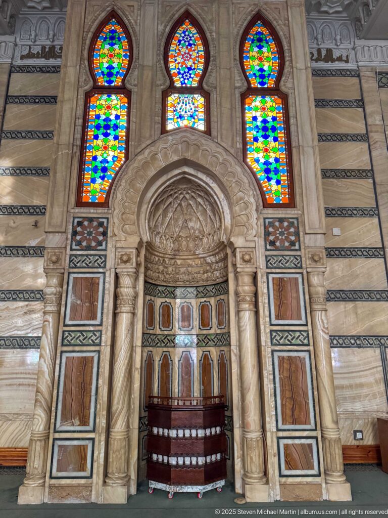 Ornate mosque prayer niche framed by carved columns and arches, with three colorful stained-glass windows above.